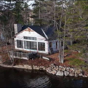 Waterfront house under construction with a black metal roof and large window openings, above an armour-stone shoreline
