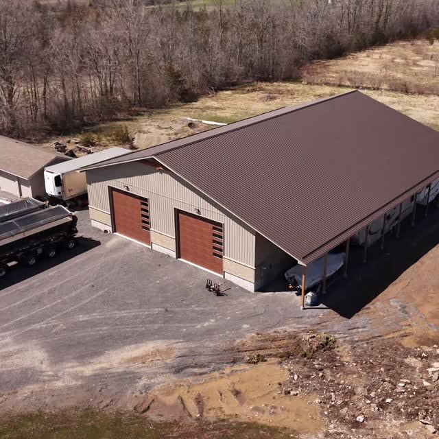 Aerial view of a purpose-built shop with a brown metal roof, two overhead doors, board-and-batten and stone exterior, and a timber-posted lean-to along one side